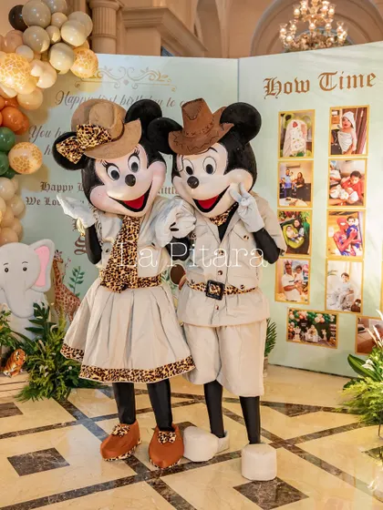 Safari-clad Mickey and Minnie mascots posing next to a milestone photo board, ready to celebrate a first birthday.
