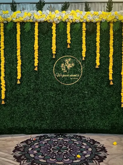 A simple photo booth or backdrop using a green hedge wall and hanging marigold garlands with bells.