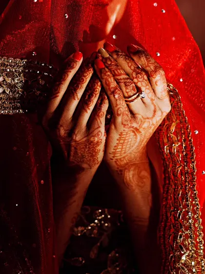 A close-up, artistic shot of a bride's henna-covered hands held in prayer, covered by a red veil.