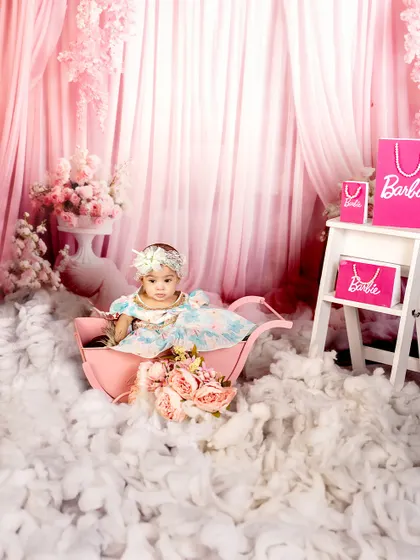 Every little princess deserves her Barbie moment. This baby girl looks serene and beautiful in a pink pram, surrounded by fluffy clouds and floral arrangements.