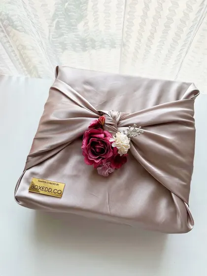 A square gift wrapped in luxurious satin fabric using the Furoshiki technique. The floral accent and branded metal tag complete the look, making it a perfect gift for Eid, weddings, or any special occasion.