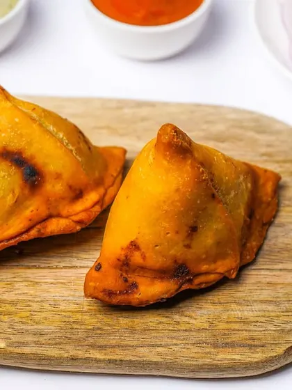 Two baked samosas with a slightly charred, crispy crust, presented simply on a wooden board. This image highlights a healthier alternative to the fried version, shot cleanly for a menu.