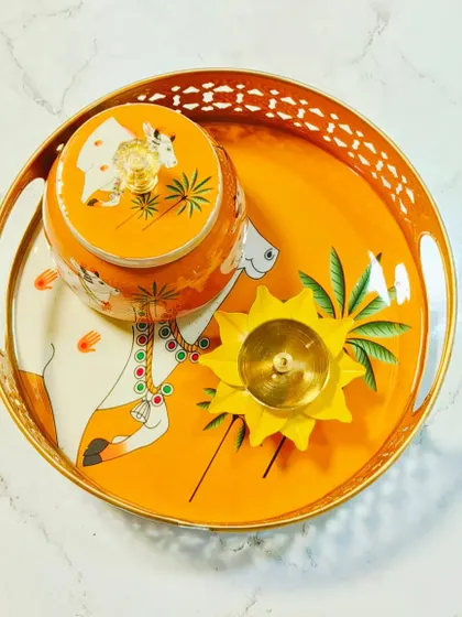 This orange Pichwai tray set features a traditional cow motif, perfect for pooja or as a decorative centerpiece. The set includes a matching jar and a brass diya, blending utility with artistic charm.