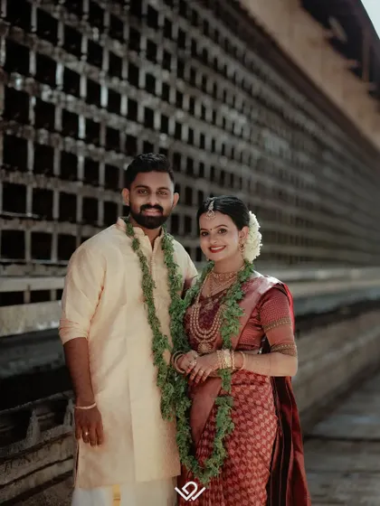A full-length portrait of a couple in their traditional South Indian wedding attire, showcasing the complete look against a simple backdrop.