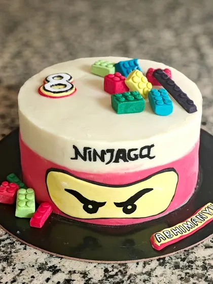 A Ninjago-themed cake with a dual-tone buttercream finish. The design features the ninja's eyes and is surrounded by colorful, edible Lego blocks made from chocolate.
