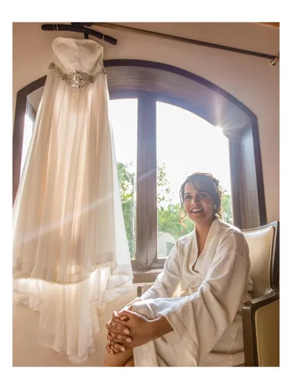 The bride smiling, sitting by the window with her wedding dress hanging beside her.
