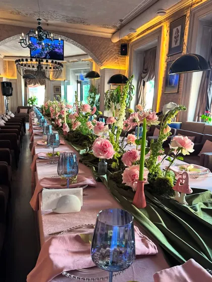 A beautifully styled long table for an intimate family gathering. The tablescape features a lush floral and moss runner, pink linens, and elegant blue glassware, creating a cozy and connected dining experience.