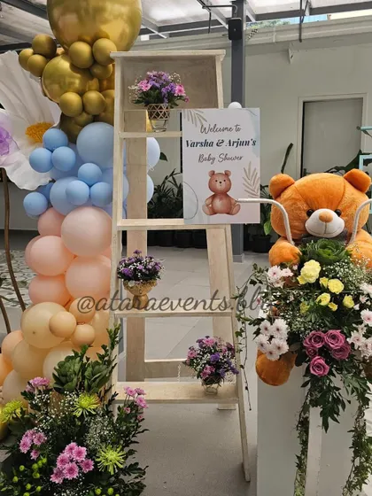 A welcome setup for a teddy bear-themed baby shower. The sign is placed on a white ladder, surrounded by flowers, balloons, and a large teddy bear, creating a sweet and inviting entrance.