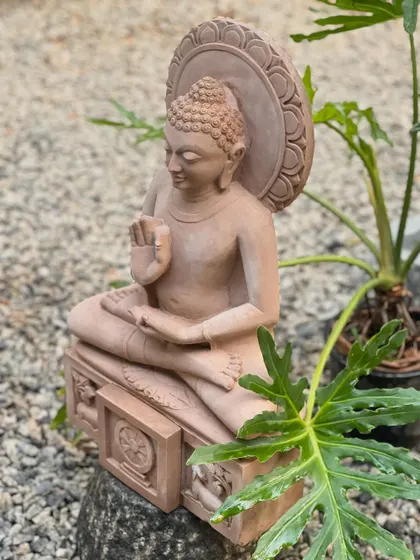 The pink sandstone Buddha statue in a garden setting. In this teaching posture, it brings a sense of serenity and wisdom, embodying peace wherever it is placed.