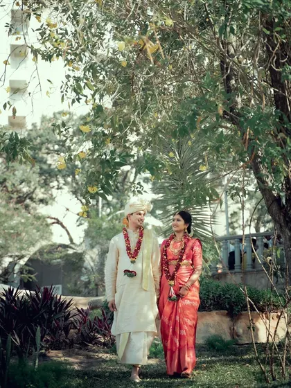 A couple in traditional wedding attire, walking hand-in-hand through a garden, sharing a happy moment.