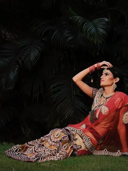 A full-length bridal portrait taken outdoors at night. The bride reclines gracefully on the grass, her red and gold lehenga spread out, creating a regal and artistic composition.