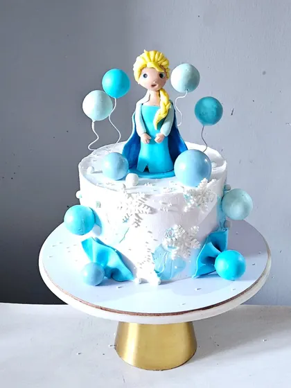 A sweet and simple cake featuring a handcrafted Elsa figurine surrounded by blue balloon-like spheres. The cake is decorated with edible snowflakes, capturing the magic of Frozen.