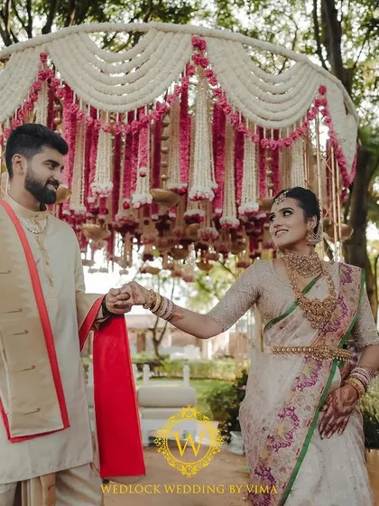 A beautiful bride standing before a traditional mandap decorated with pink and white floral tassels. The hanging brass urlis and rose drops add a touch of classic elegance to the South Indian ceremony.