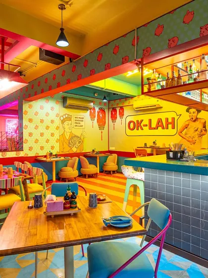 This view of The Fatty Bao shows the interplay between different sections of the restaurant. The "OK-LAH" wall art and varied seating arrangements are framed to showcase the dynamic and multi-layered design of the space.