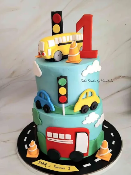 A colourful two-tier first birthday cake with a car and bus theme. The design features a road, traffic lights, and cute vehicles, perfect for a little transport fan.