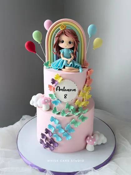 A beautiful two-tiered cake with a butterfly and rainbow theme. This pink cake is adorned with a cascade of colorful, edible butterflies and a handcrafted doll.