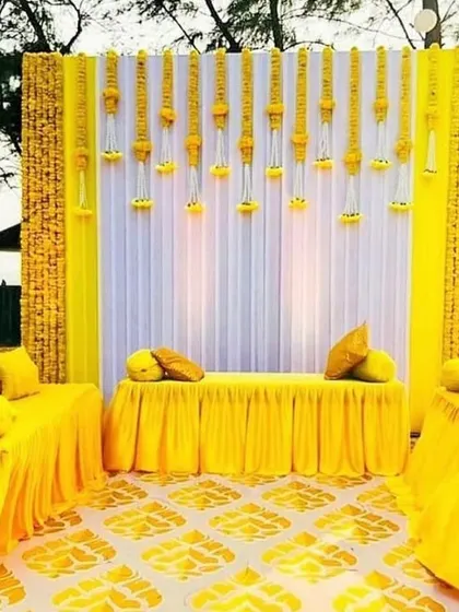 A bright and sunny Haldi ceremony setup with a yellow and white color scheme. The backdrop features hanging floral strings made of marigolds, creating a traditional and festive atmosphere.