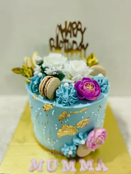 A beautiful blue cake for "Mumma," adorned with gold leaf, macarons, and a mix of white, blue, and purple flowers.