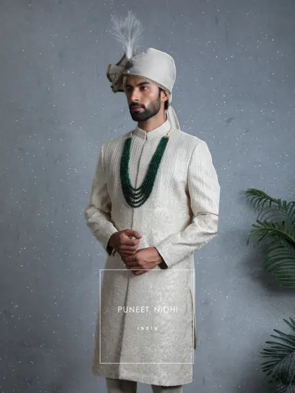 An ivory sherwani sculpted with intricate artistry, crowned with a feathered turban and an emerald mala. It embodies the quiet strength and dignity every groom carries into his new chapter.