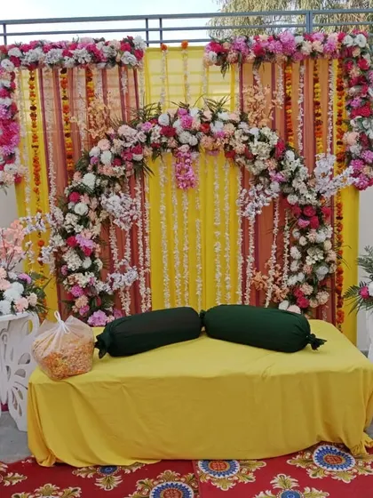 A floral-heavy backdrop for a pre-wedding ceremony, featuring a dense arch of artificial flowers over a yellow draped background.
