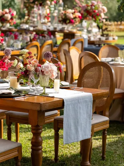 A close-up of a tablescape at an outdoor anniversary lunch. The wooden table, cane-back chairs, and simple runner create a rustic yet refined look.