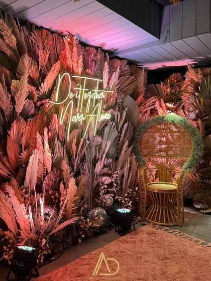 A dramatic and moody photo booth with a wall of dried pampas and palm leaves, illuminated by a neon sign. The wicker peacock chair adds the perfect finishing touch.