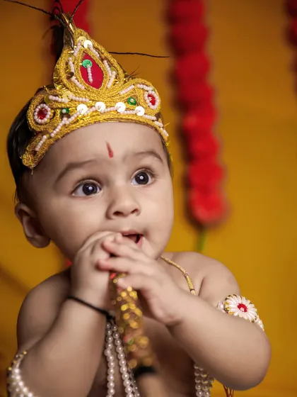 A beautiful close-up of a baby Krishna with wide, curious eyes. The soft lighting and warm yellow background highlight his innocent features.