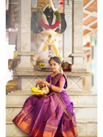 A young girl in a traditional purple saree sits on the steps of a temple, holding a basket of offerings for a cultural photoshoot.