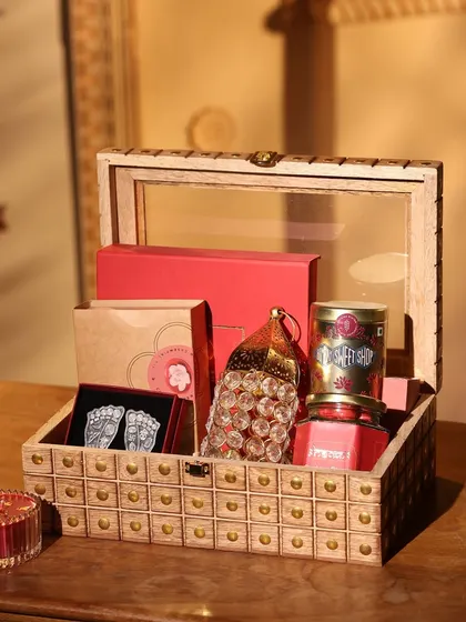 A classic wooden box with a glass lid, filled with red-themed gifts for Diwali. This hamper includes a stunning crystal lantern, silver-plated Lakshmi feet, and gourmet sweets.