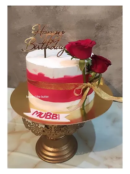 A classic red and white birthday cake. The simple design is elevated by a red ribbon, fresh red roses, and a gold "Happy Birthday" topper.
