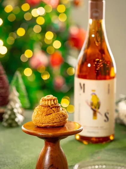 A festive shot of a Christmas pastry on a wooden stand, with a bottle of wine and a decorated tree in the background.