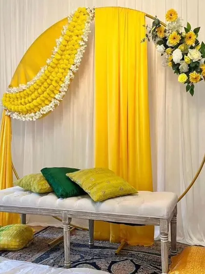 A simple and modern Haldi seating area with a circular golden frame. The frame is decorated with yellow drapes and a partial floral arrangement, creating a chic and minimalist look.