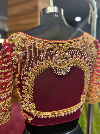This maroon bridal blouse is a work of art, featuring a heart-shaped cutout on the back. The design is framed with intricate zardosi and pearl work, with delicate beaded tassels adding movement and charm.