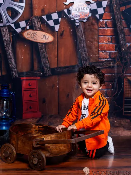 A happy toddler playing with a wooden wagon in our rustic garage-themed studio. The warm lighting and detailed props create a lively and engaging scene for a boy's photoshoot.