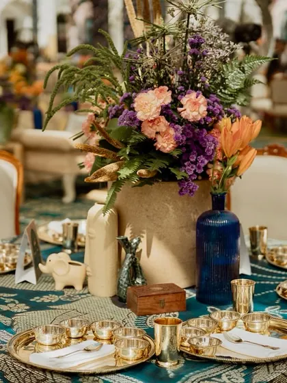 An intimate dining table set with handcrafted brass thalis for an authentic Rajasthani meal. The table is adorned with a hand-blocked print tablecloth and a centerpiece of jewel-toned flowers.