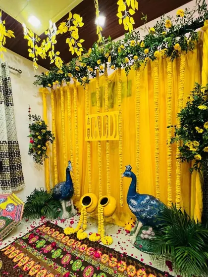 A beautiful Haldi decoration setup for a home function. The decor features a yellow drape backdrop, peacock statues, and a colorful traditional rug.