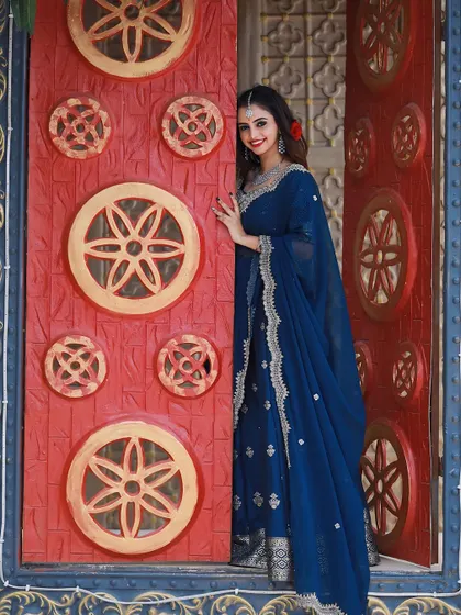 The contrast of the royal blue lehenga against the traditional red door makes for a stunning visual.
