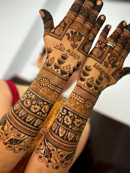 This full-arm bridal design is rich with detail, including traditional floral bands, elephant motifs, and sacred diyas. The names of the bride and groom are elegantly written in Hindi, adding a personal touch.