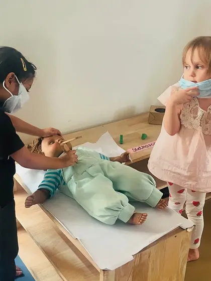 Two children play doctor, using masks and a toy thermometer to care for a baby doll. This helps them process their own experiences with doctors in a safe, controlled way.