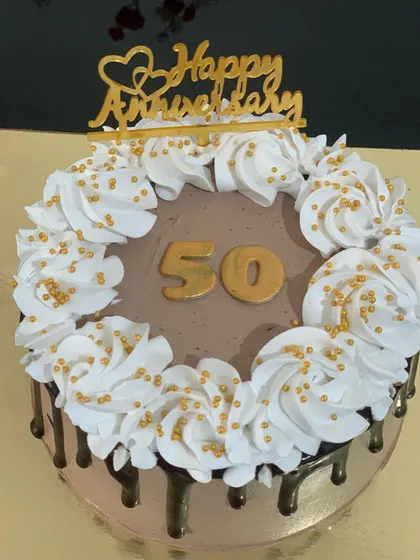 A beautiful cake for a 50th anniversary or birthday. The chocolate cake has a dark chocolate drip and is topped with white cream swirls and gold sprinkles.