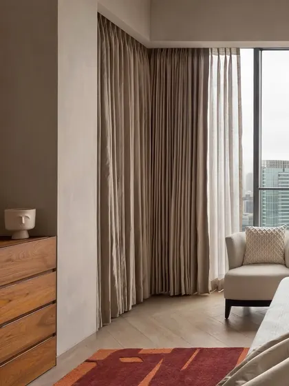 This corner of the East Coast Residence bedroom shows the simple, elegant lines of the wooden dresser and the soft, floor-to-ceiling curtains that frame the city view.