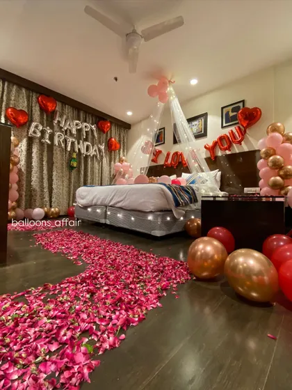 A romantic birthday surprise filling a hotel room with love. The decor includes a rose petal pathway, a canopy with fairy lights over the bed, 'I Love You' and 'Happy Birthday' foil balloons, and balloon arrangements in red and rose gold.