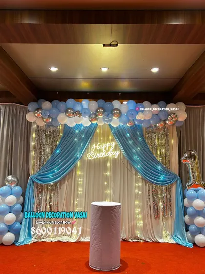 A grand stage decoration for a first birthday. The setup features blue and white drapes, a shimmer curtain with fairy lights, and a neon sign.