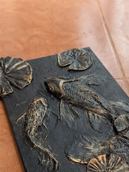 An angled view of the finished koi fish textured art. The metallic gold paint catches the light differently across the raised surfaces, creating a sense of movement and depth.