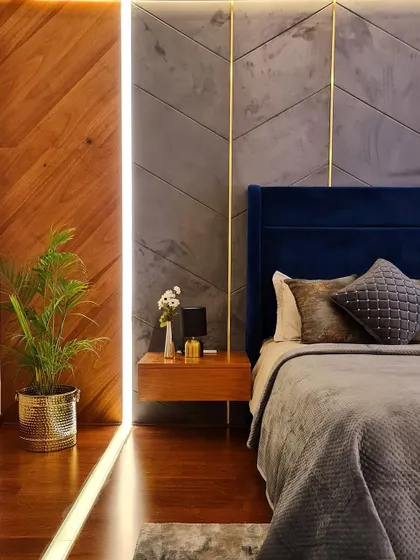 A master bedroom designed with rich, warm tones. A deep blue velvet headboard contrasts beautifully with a grey textured wall featuring brass inlays. The floating wood nightstand and integrated lighting add to the modern, luxurious feel.