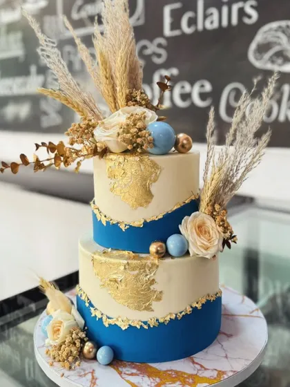 A modern two-tiered cake perfect for a stylish celebration. This design combines a rich royal blue base with an ivory top tier, accented with edible gold leaf, dried pampas grass, and delicate sugar flowers.
