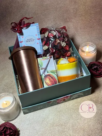 Shine bright with our Diwali delights. This hamper includes a copper tumbler, a layered candle, a bag of potpourri, and a box of chocolates, all packed in a beautiful floral gift box.