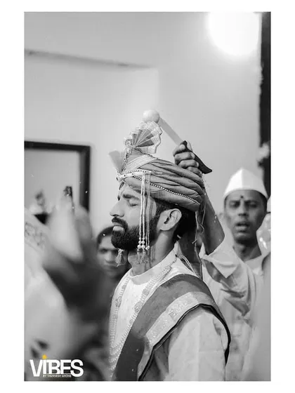 A black and white shot capturing the groom during a pre-ceremony ritual, as his turban is being adjusted.