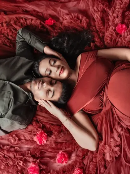 An artistic and intimate top-down shot of an expecting couple. Lying amidst a bed of red fabric and petals, this photo captures a moment of quiet connection and love.