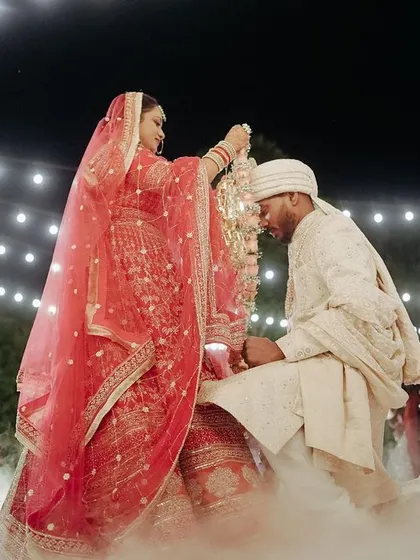 The bride places the Varmala on her groom, who is kneeling, in a beautiful, smoke-filled, nighttime ceremony.